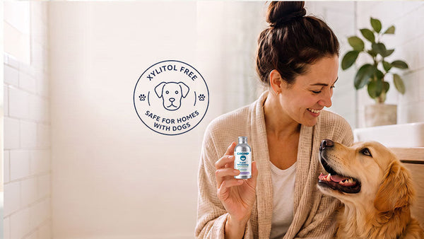 A woman in a beige robe holds a small bottle for her golden retriever dog in a bright bathroom. A circular badge on the left reads 'Xylitol Free Safe for Homes with Dogs'.