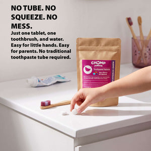 A hand is shown placing a white round tablet on a white bathroom counter next to a stand-up pouch of Chomp Junior toothpaste tablets. The scene includes a toothbrush, a tube of toothpaste, and a toothbrush holder in the background, with promotional text on the left.