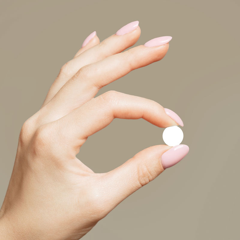 A close-up of a hand with pale pink manicured nails holding a small, round white pill between the fingers against a solid taupe background.