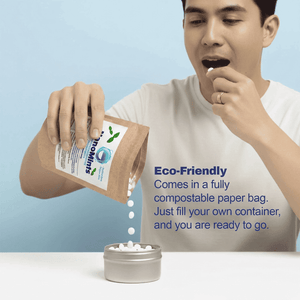 A man in a white t-shirt is shown pouring white tablets from a brown paper pouch labeled 'NanoMints' into a small silver metal container. The background is a solid light blue, with promotional text on the right describing the eco-friendly compostable packaging.