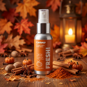 A bottle of CHOMP Pumpkin Spice Fresh Spray stands on a wooden surface surrounded by autumn spices, cinnamon sticks, star anise, small pumpkins, and scattered pumpkin spice powder. The background features blurred fall leaves and a warm glowing lantern.