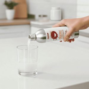 Hand pouring a liquid from a silver bottle labeled 'Chomp' into a glass on a kitchen counter.