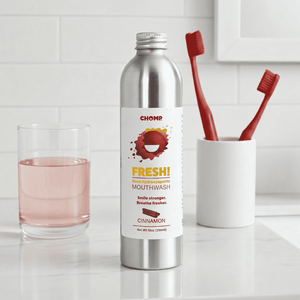 Chomp mouthwash bottle with a glass of water and toothbrushes on a bathroom counter.