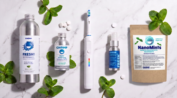 A flat-lay arrangement of Chomp oral care products on a white marble background with mint leaves. The products include a silver bottle of Fresh! Natural Mint flavor, a small clear bottle of Chomp Water Flosser, a white electric toothbrush with a rainbow accent, a small blue bottle of NanoMints, and a brown pouch of NanoMints.