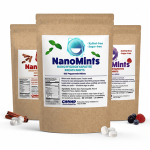 Three stand-up kraft paper pouches of NanoMints breath mint are arranged on a white studio background, with loose mint tablets and small fruit/cinnamon props at the base. The center pouch is fully visible and labeled NanoMints peppermint; the two side pouches are partially cropped and show different flavor labels.
