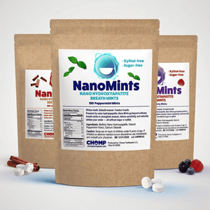 Three stand-up kraft paper pouches of NanoMints breath mint are arranged on a white studio background, with loose mint tablets and small fruit/cinnamon props at the base. The center pouch is fully visible and labeled NanoMints peppermint; the two side pouches are partially cropped and show different flavor labels.