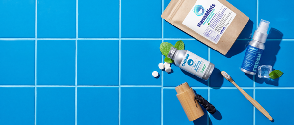 A flat-lay arrangement of dental oral care products on a bright blue tiled background, including a brown pouch labeled NanoMats, a silver bottle, a blue spray bottle, a wooden toothbrush, mint leaves, and a small wooden container.