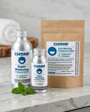 Chomp Super Whitening Toothpaste Tablets in a silver bottle and a smaller silver bottle, alongside a brown stand-up pouch of the same product, set on a white marble surface with mint leaves in the foreground.