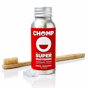 A bottle of CHOMP Super Whitening Toothpaste Tablets in Sinful Cinnamon flavor, shown with two white tablets and a bamboo toothbrush on a reflective white surface.