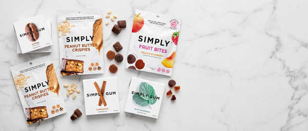 Five Simply Gum snack pouches and individual packs arranged on a white marble surface, with scattered gum pieces and small food pieces around them. Visible flavors include Peanut Butter Crispies, Fruit Bites, and Cinnamon.
