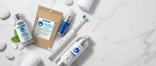 A flat-lay arrangement of Chomp oral care products on a white marble background, including a silver bottle, a kraft pouch, a blue spray bottle, a white electric toothbrush, and a silver bottle with a white cap, surrounded by white stones, a green leaf, and rolled white towels.