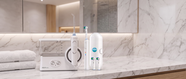 A dental oral care set on a white marble bathroom counter, featuring a water flosser, an electric toothbrush, and a white cylindrical device with a blue circular logo, set against a marble backsplash &mirror.