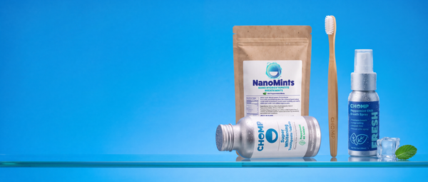 A dental care product set on a glass shelf against a bright blue background, featuring a brown stand-up pouch labeled NanoMints Breath Mints, a silver bottle labeled CHOMP Super Whitening Toothpaste Tablets, a blue CHOMP Fresh Breath Spray bottle, a wooden toothbrush, mint leaves, and a few white tablets.