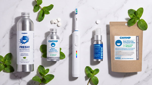 A flat-lay arrangement of Chomp oral care products on a white marble background with mint leaves. The products include a silver bottle of Fresh! mouthwash, a small bottle of toothpaste, a white electric toothbrush, a blue spray bottle, and a brown pouch of peppermint gum.