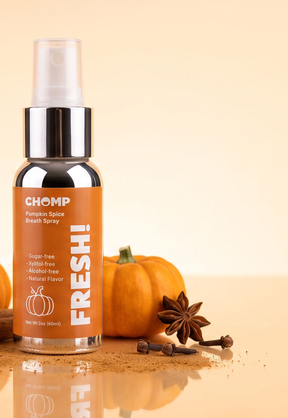 A bottle of CHOMP Pumpkin Spice Breath Spray stands on a reflective surface with a small orange pumpkin, star anise, and cloves beside it against a warm beige background.