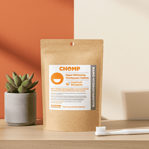 CHOMP toothpaste tablets packaging on a wooden surface with a plant and toothbrush in the background.