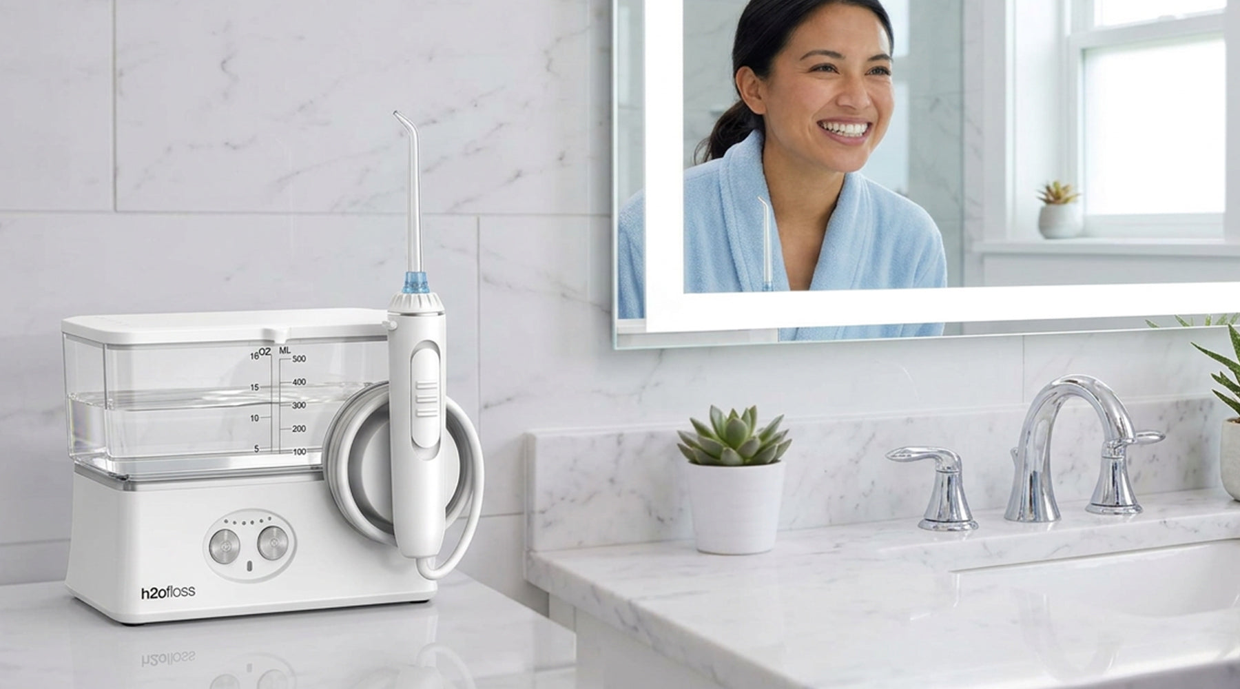 A white oral irrigator water flosser and its charging base sit on a bathroom counter beside a mirror showing a smiling woman in a blue robe. The bathroom has white marble surfaces and a chrome faucet.