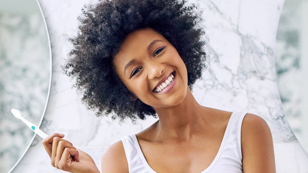 A smiling woman with an afro sits on a white bathroom vanity, wearing a white tank top and denim shorts while holding a toothbrush. The background features a white and gray marble wall with two large round mirrors.
